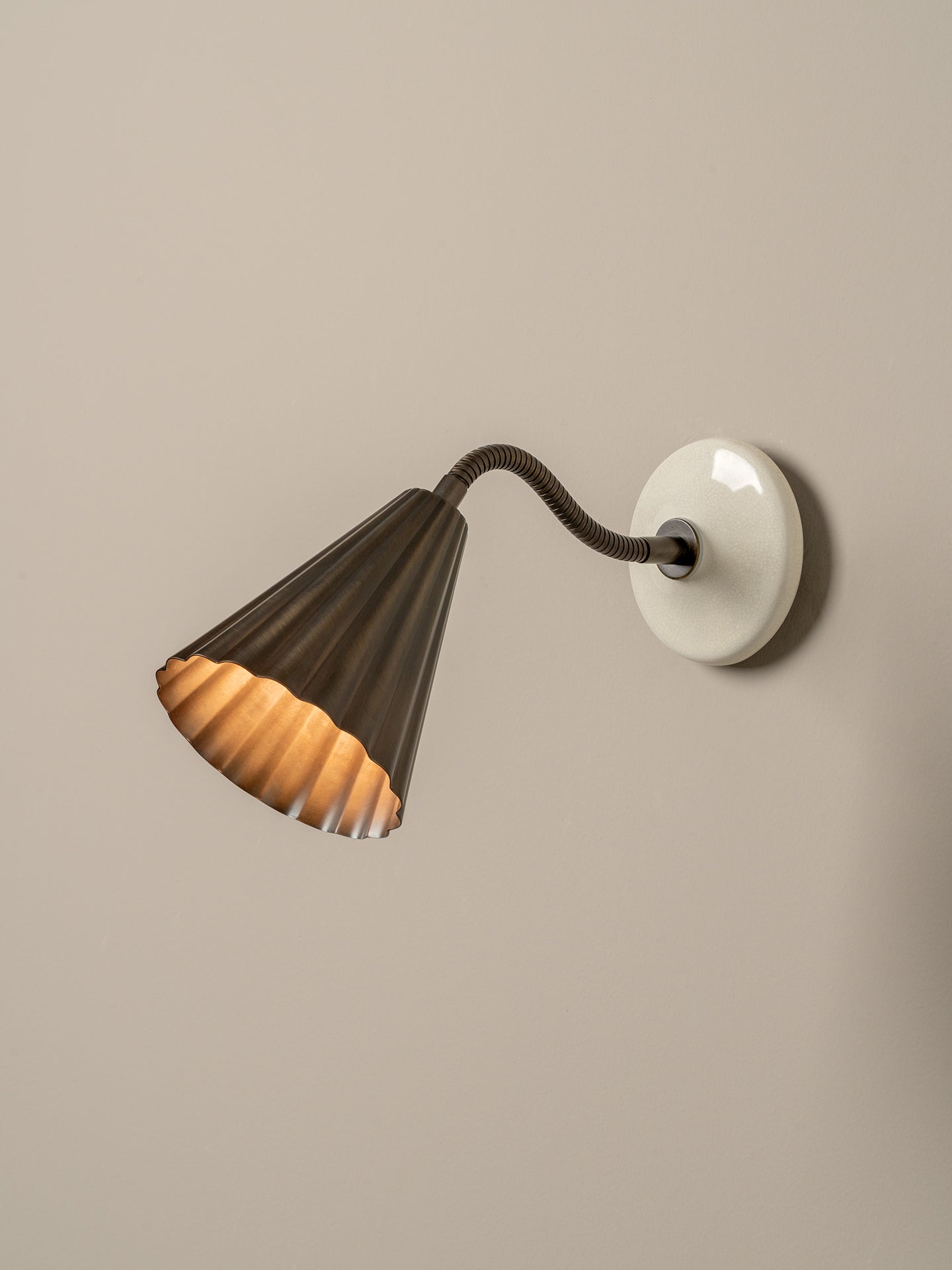 A modern wall sconce with a dark bronze fluted shade, a flexible dark metal gooseneck, and a round white wall mount, shown lit against a neutral beige wall.