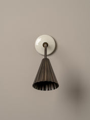 A modern wall-mounted sconce with a fluted dark metal shade and a round white wall plate, shown against a plain warm gray wall.