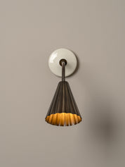 A modern wall-mounted light fixture with a white round backplate, a dark metal gooseneck, and a pleated conical shade that glows warm yellow from within, set against a solid taupe background.