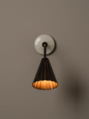A modern wall sconce with a black pleated conical shade and a warm glowing orange interior, mounted on a round white wall plate against a solid taupe background.