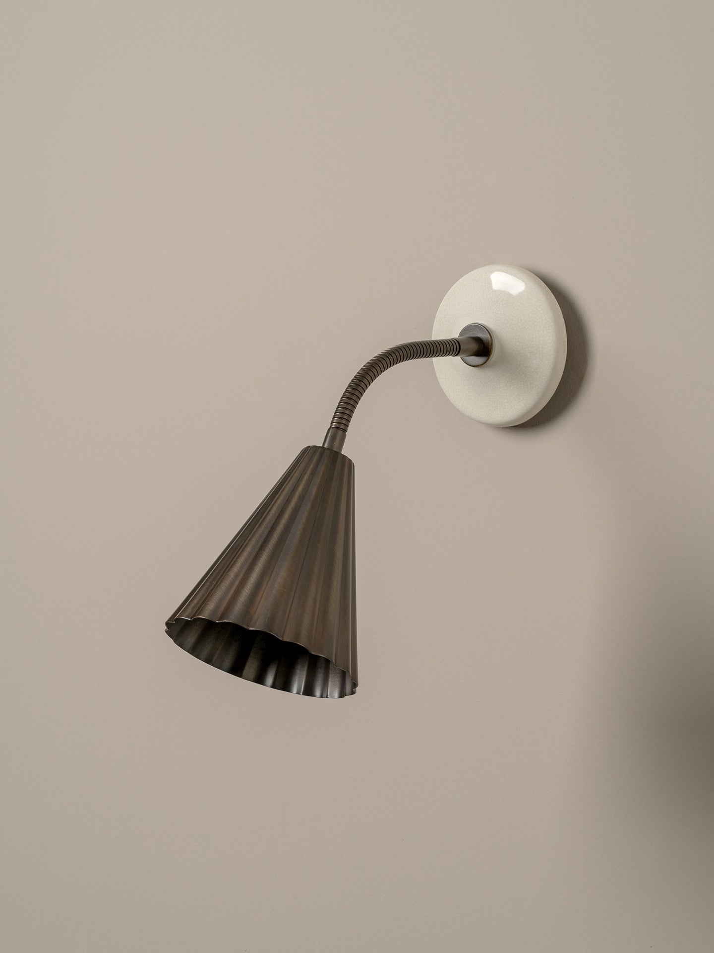 A modern wall-mounted sconce with a dark bronze fluted shade, a flexible curved neck, and a round white wall plate, shown against a plain beige wall.
