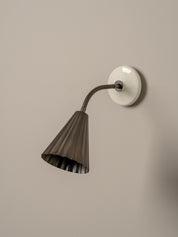 A modern wall-mounted sconce with a dark bronze fluted shade, a flexible curved neck, and a round white wall plate, shown against a plain beige wall.