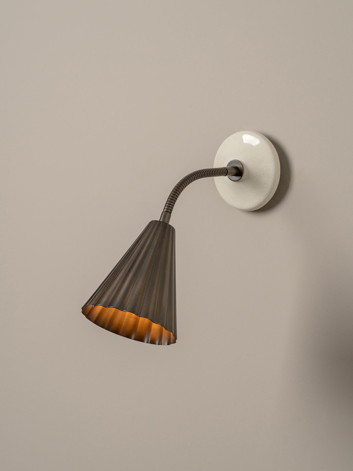A modern wall sconce with a dark gray pleated metal shade, a flexible gooseneck arm, and a round white wall mount, shown against a neutral beige wall.