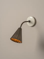 A modern wall sconce with a dark gray pleated metal shade, a flexible gooseneck arm, and a round white wall mount, shown against a neutral beige wall.