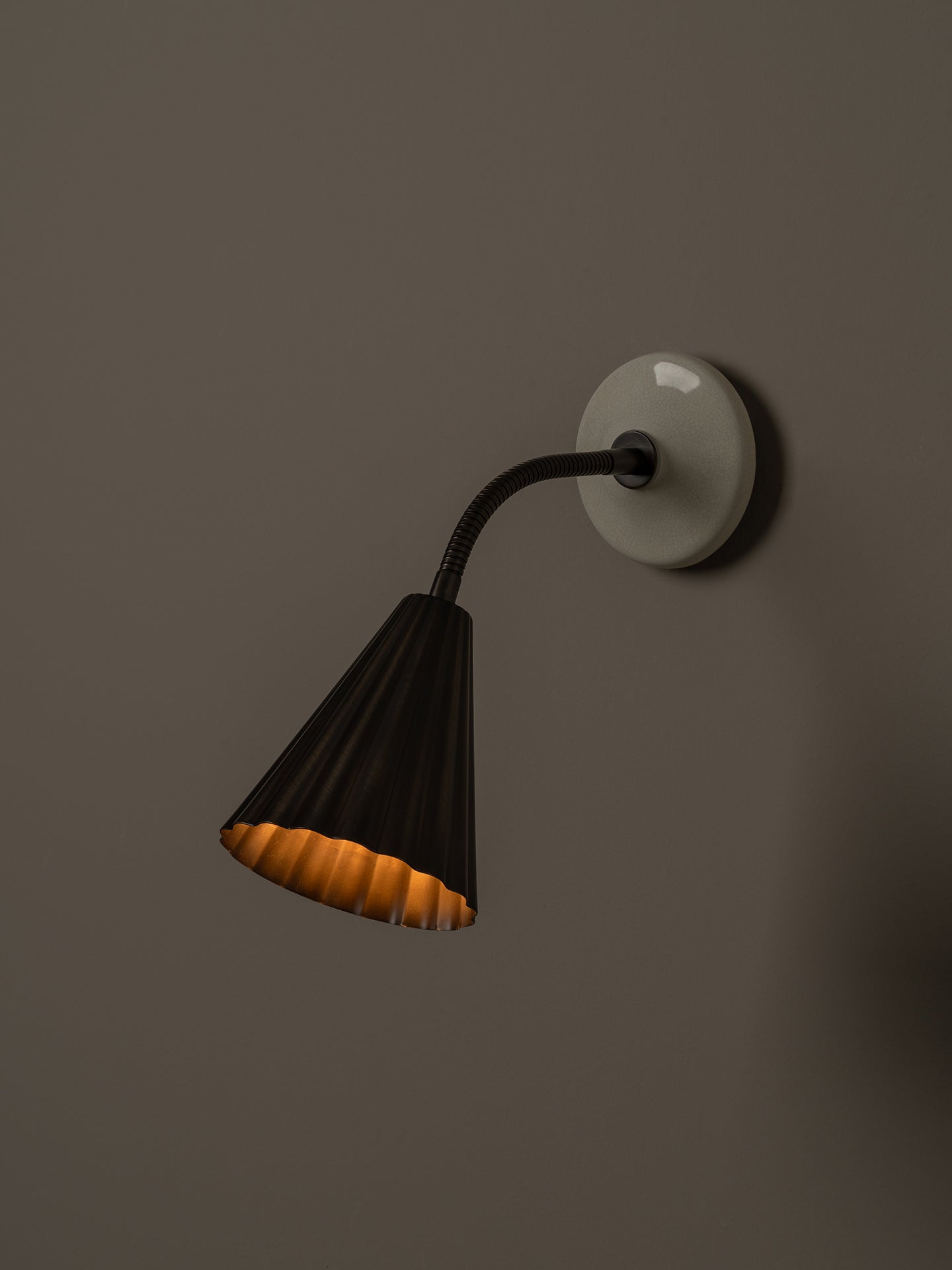 A modern wall-mounted sconce with a black pleated shade, black flexible gooseneck arm, and a round off-white wall plate, shown lit against a dark brown wall.