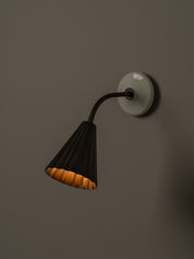 A modern wall-mounted sconce with a black pleated shade, black flexible gooseneck arm, and a round off-white wall plate, shown lit against a dark brown wall.