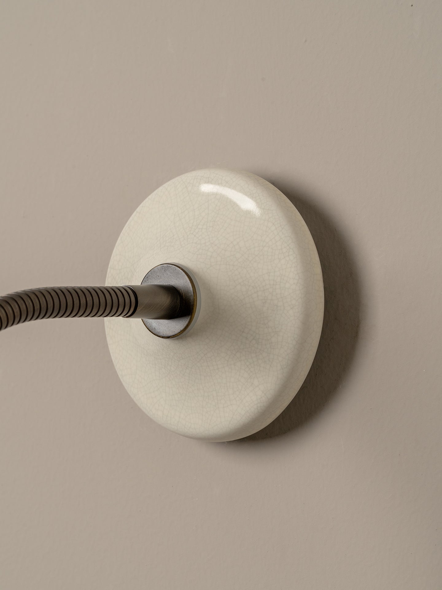 A minimalist wall-mounted light fixture with a round, glossy off-white ceramic or glass shade and a dark gray flexible arm, set against a solid taupe background.