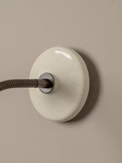 A minimalist wall-mounted light fixture with a round, glossy off-white ceramic or glass shade and a dark gray flexible arm, set against a solid taupe background.
