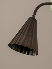A close-up of a dark bronze flexible gooseneck lamp shade with a fluted, scalloped edge, set against a solid warm beige background.