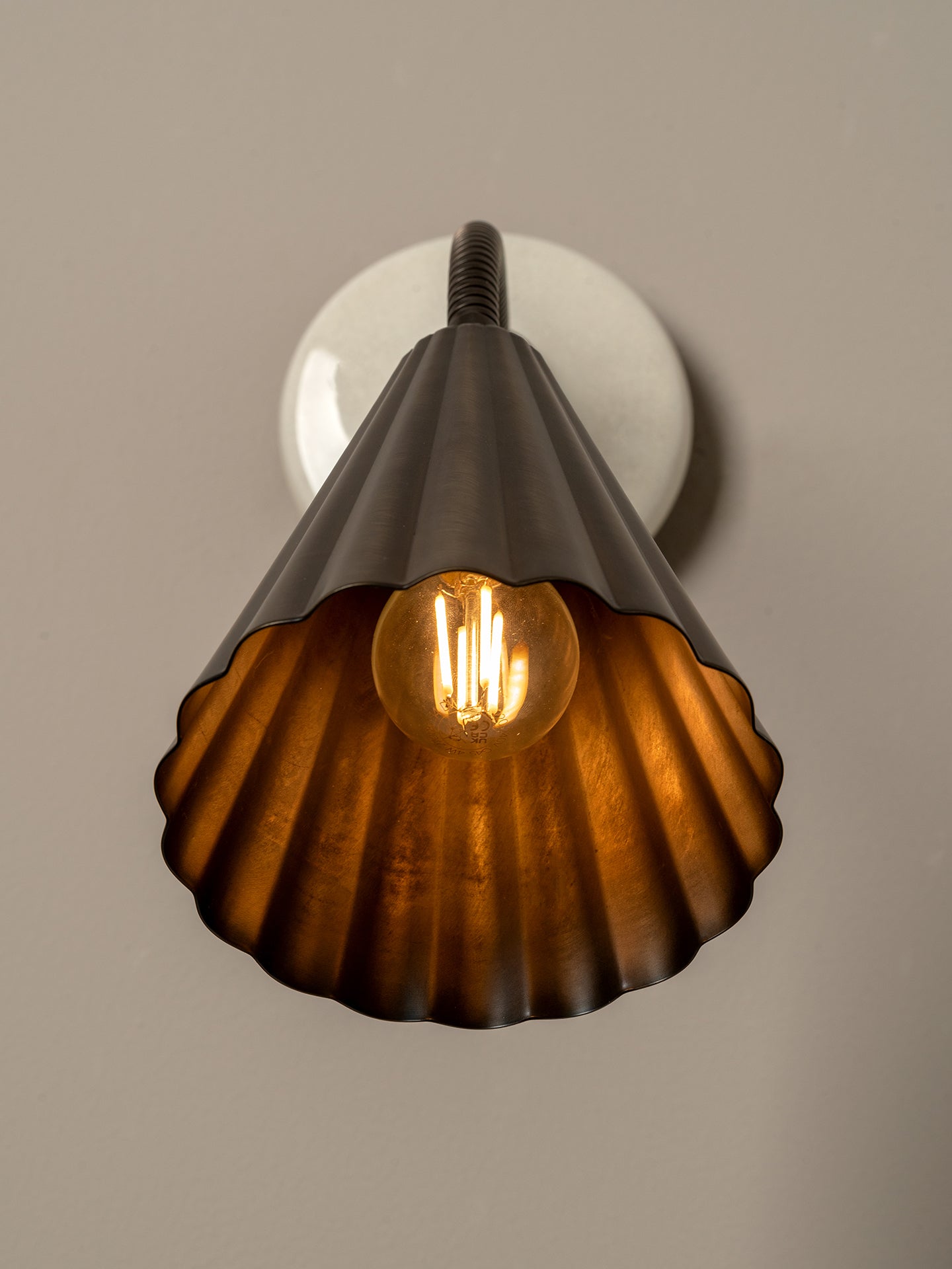 A vintage-style industrial wall sconce featuring a ribbed, scalloped metal shade in a dark bronze finish, illuminated by a glowing warm Edison bulb. The fixture is mounted on a round white wall plate against a neutral, warm gray-beige background.