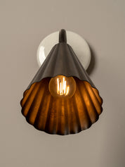 A vintage-style industrial wall sconce featuring a ribbed, scalloped metal shade in a dark bronze finish, illuminated by a glowing warm Edison bulb. The fixture is mounted on a round white wall plate against a neutral, warm gray-beige background.