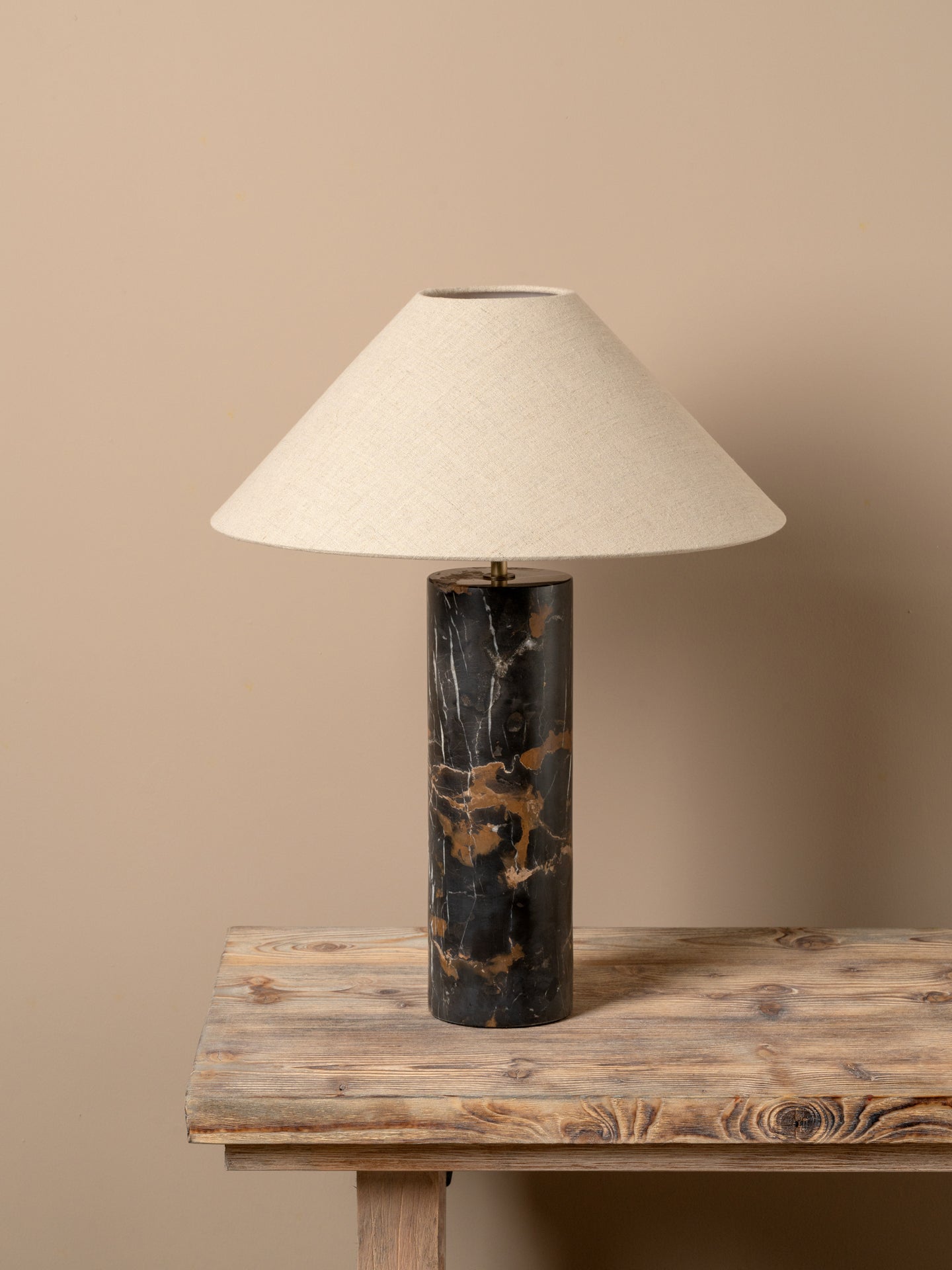 A table lamp with a tapered off-white fabric shade and a cylindrical black marble base with gold veining, displayed on a rustic wooden table against a solid beige background.
