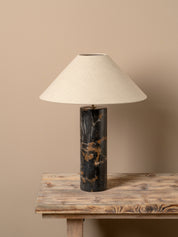 A table lamp with a tapered off-white fabric shade and a cylindrical black marble base with gold veining, displayed on a rustic wooden table against a solid beige background.