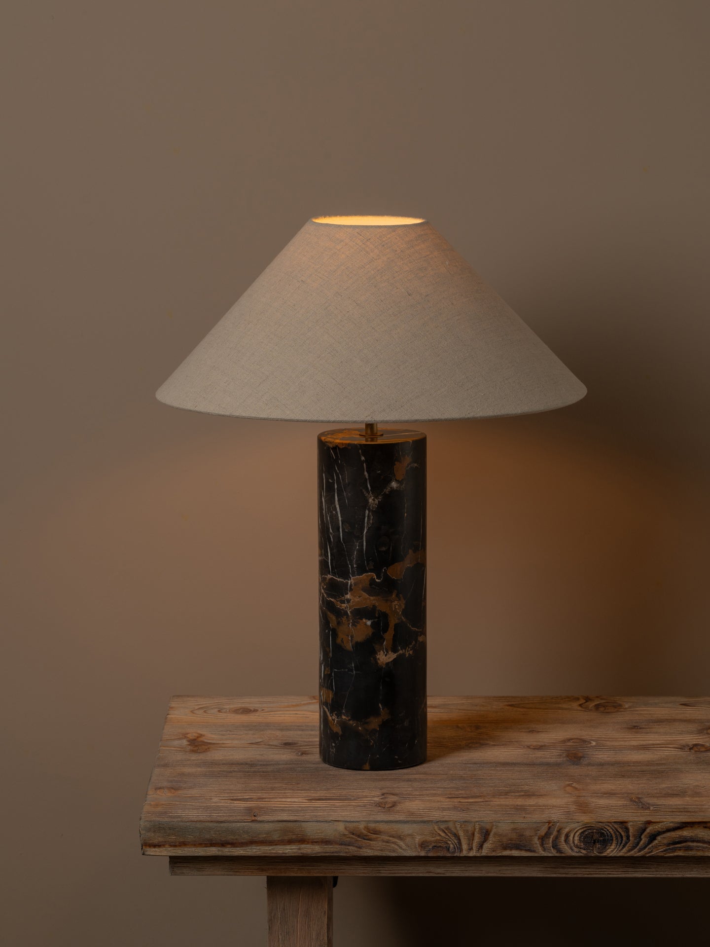 A table lamp with a tapered off-white fabric shade and a cylindrical black marble base with gold veining, shown lit on a rustic wooden table against a warm brown background.
