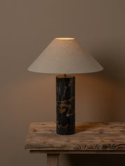 A table lamp with a tapered off-white fabric shade and a cylindrical black marble base with gold veining, shown lit on a rustic wooden table against a warm brown background.