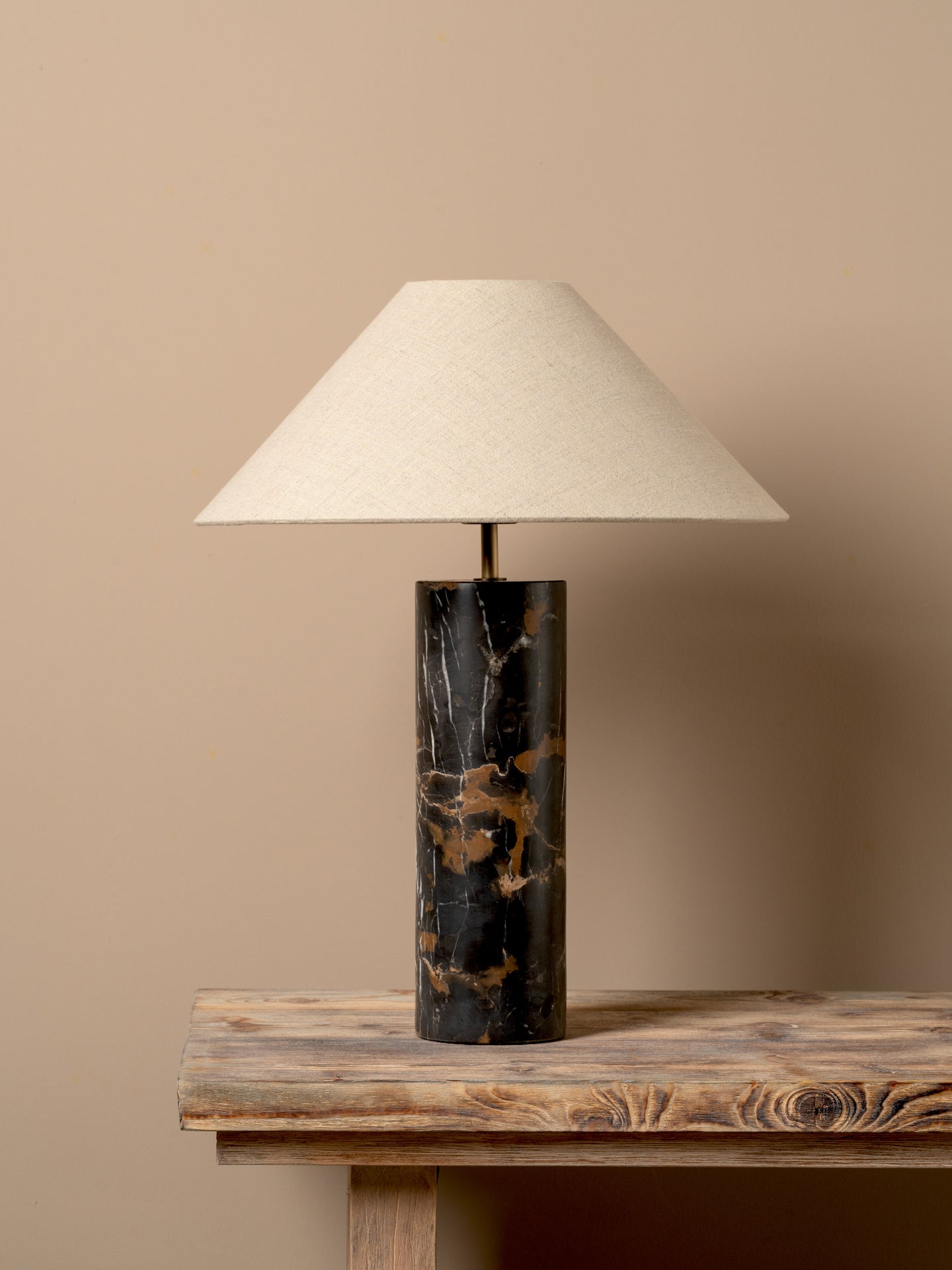 A table lamp with a tapered off-white fabric shade and a cylindrical black marble base featuring natural gold veining, displayed on a rustic wooden table against a solid beige background.