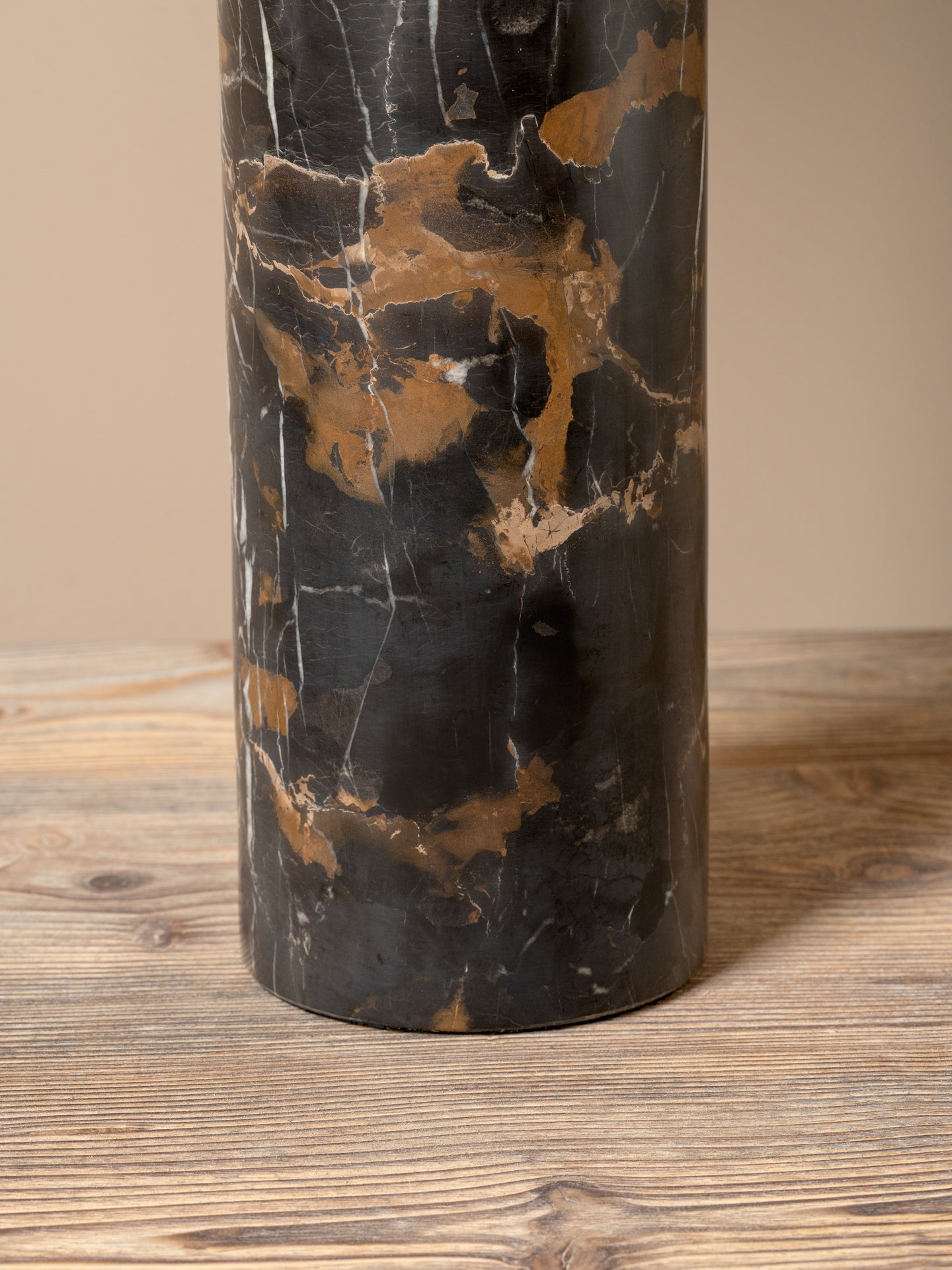 A cylindrical table lamp base made of polished black and bronze-brown marble, standing on a rustic wooden surface against a solid beige background.