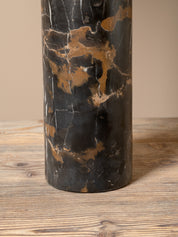 A cylindrical table lamp base made of polished black and bronze-brown marble, standing on a rustic wooden surface against a solid beige background.
