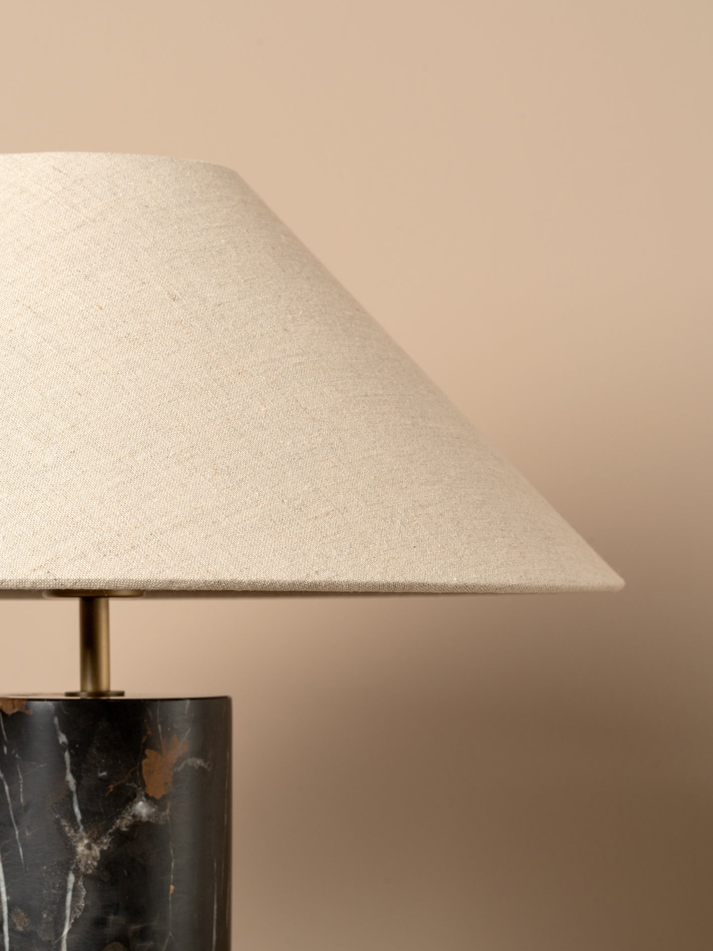 Close-up of a table lamp with a tapered off-white linen shade and a dark marble base with gold veining, set against a solid beige background.