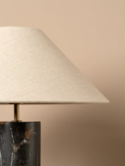 Close-up of a table lamp with a tapered off-white linen shade and a dark marble base with gold veining, set against a solid beige background.