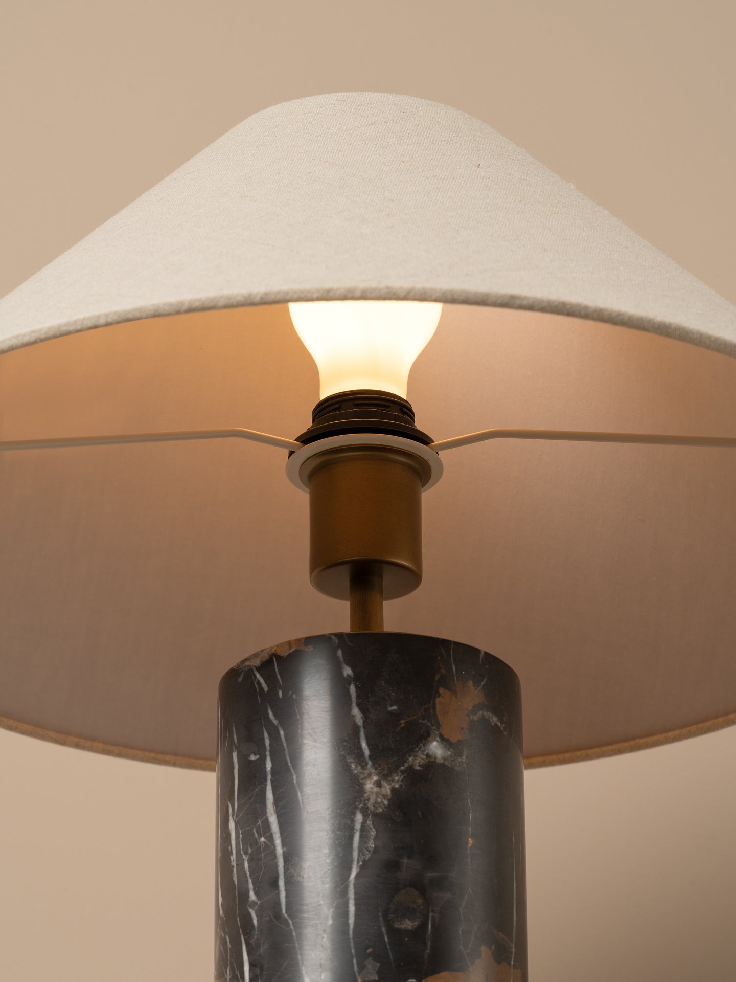 Close-up of a table lamp with a white fabric drum shade, a lit bulb, a brass socket, and a dark marble cylindrical base against a warm beige background.