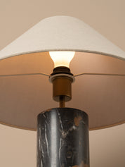 Close-up of a table lamp with a white fabric drum shade, a lit bulb, a brass socket, and a dark marble cylindrical base against a warm beige background.