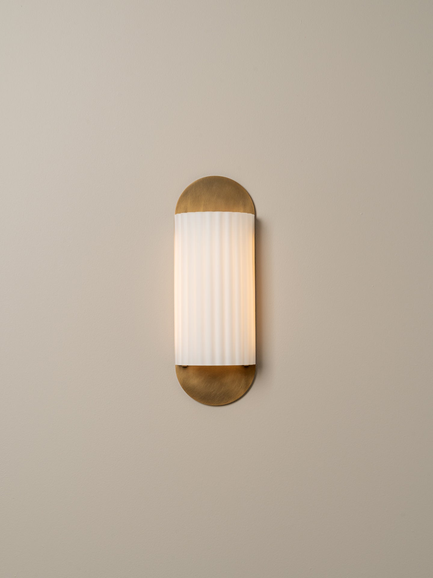A modern wall sconce light fixture with a vertical white ribbed glass shade and brushed brass metal ends, mounted on a plain beige wall.
