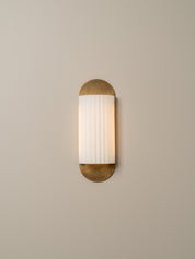 A modern wall sconce light fixture with a vertical white ribbed glass shade and brushed brass metal ends, mounted on a plain beige wall.
