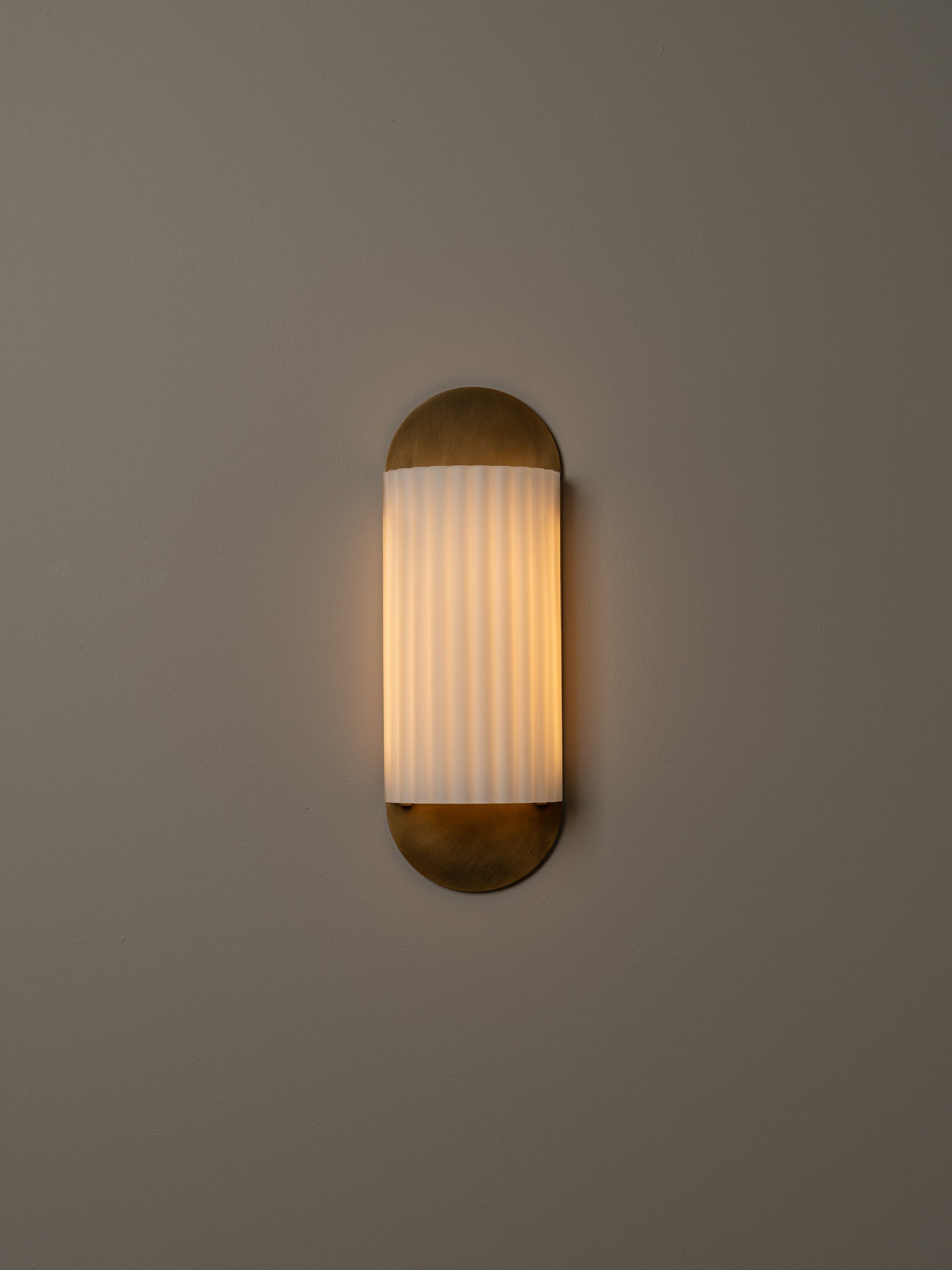 A modern wall-mounted sconce with a vertical ribbed frosted glass shade and a brass-toned metal base and top cap, shown lit against a plain warm gray wall.