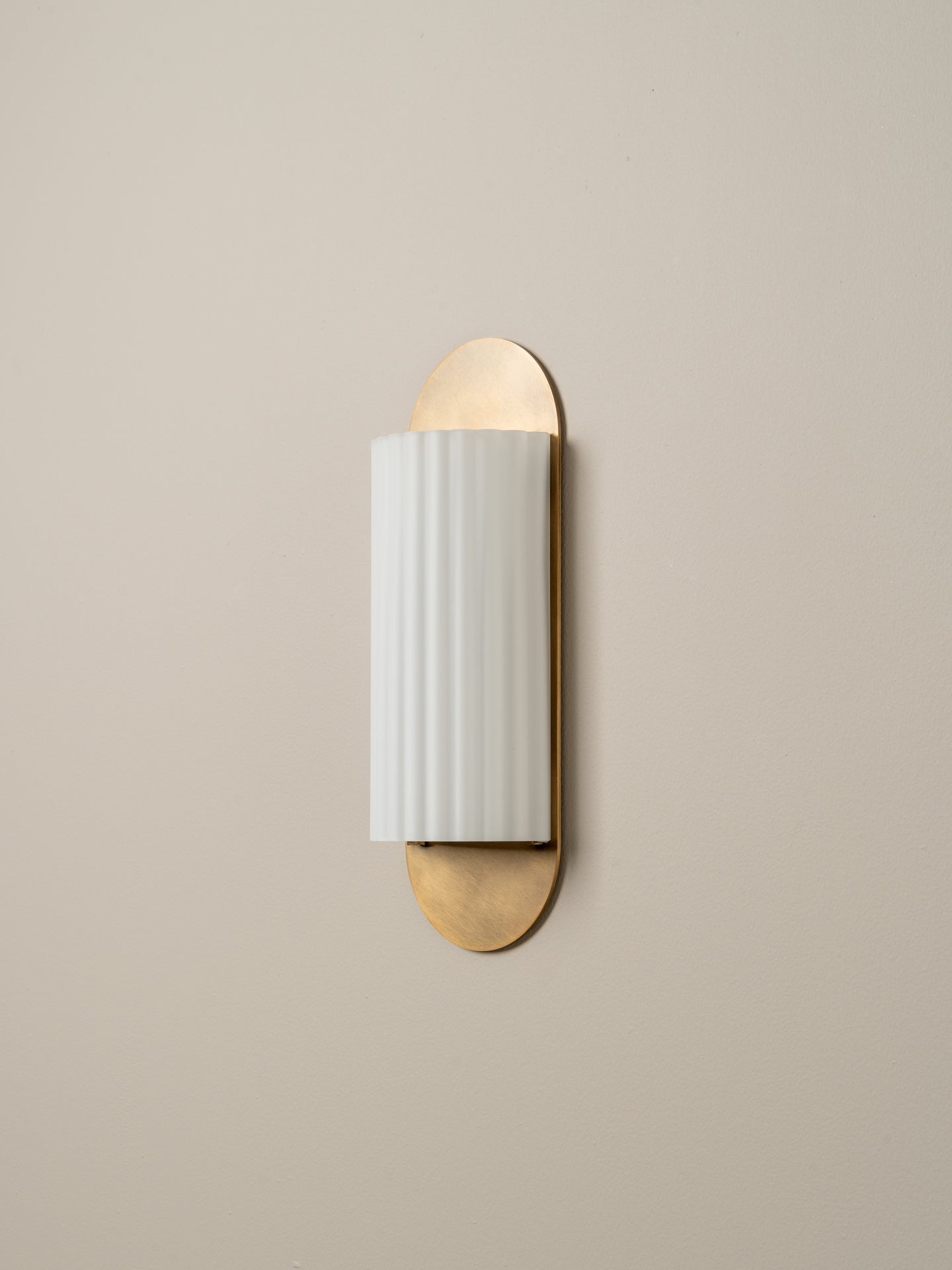 A modern wall-mounted sconce with a vertical white ribbed glass shade and brushed brass metal accents on a warm beige wall.