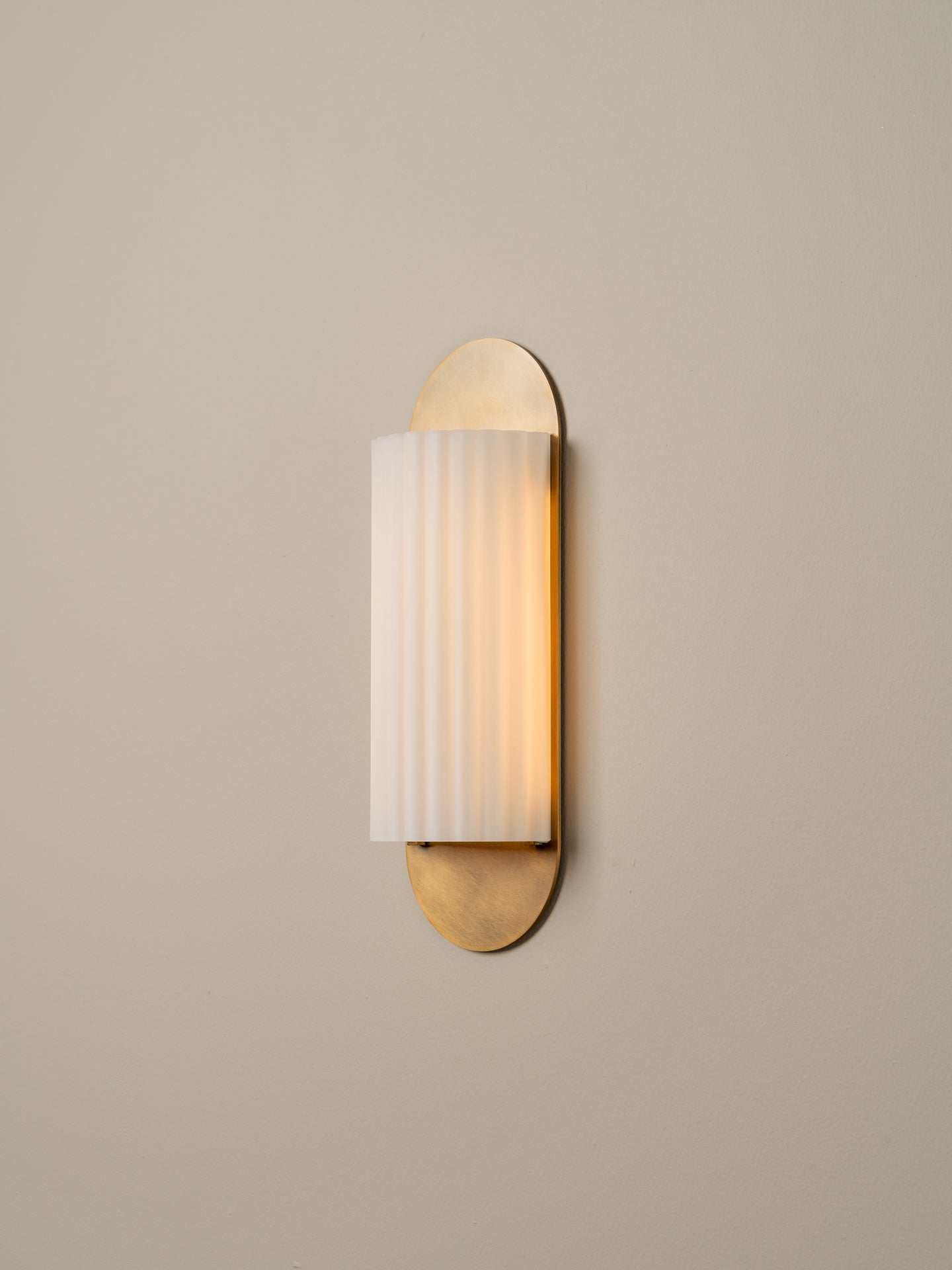 A modern wall sconce light fixture with a vertical frosted glass shade featuring subtle vertical ridges, flanked by two oval brushed brass metal caps, mounted on a plain beige wall.