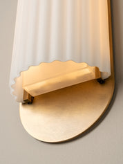 Close-up of a wall-mounted light fixture with a ribbed frosted glass shade and a brushed gold metal base, shown against a plain light wall.