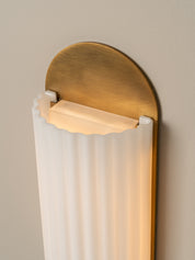 A modern wall sconce light fixture with a brushed brass semi-circular backplate, a white ribbed glass shade, and a warm glowing light, mounted on a beige wall.