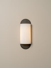 A modern wall sconce light fixture with a vertical ribbed white glass shade and dark charcoal metal caps, mounted on a plain beige wall.
