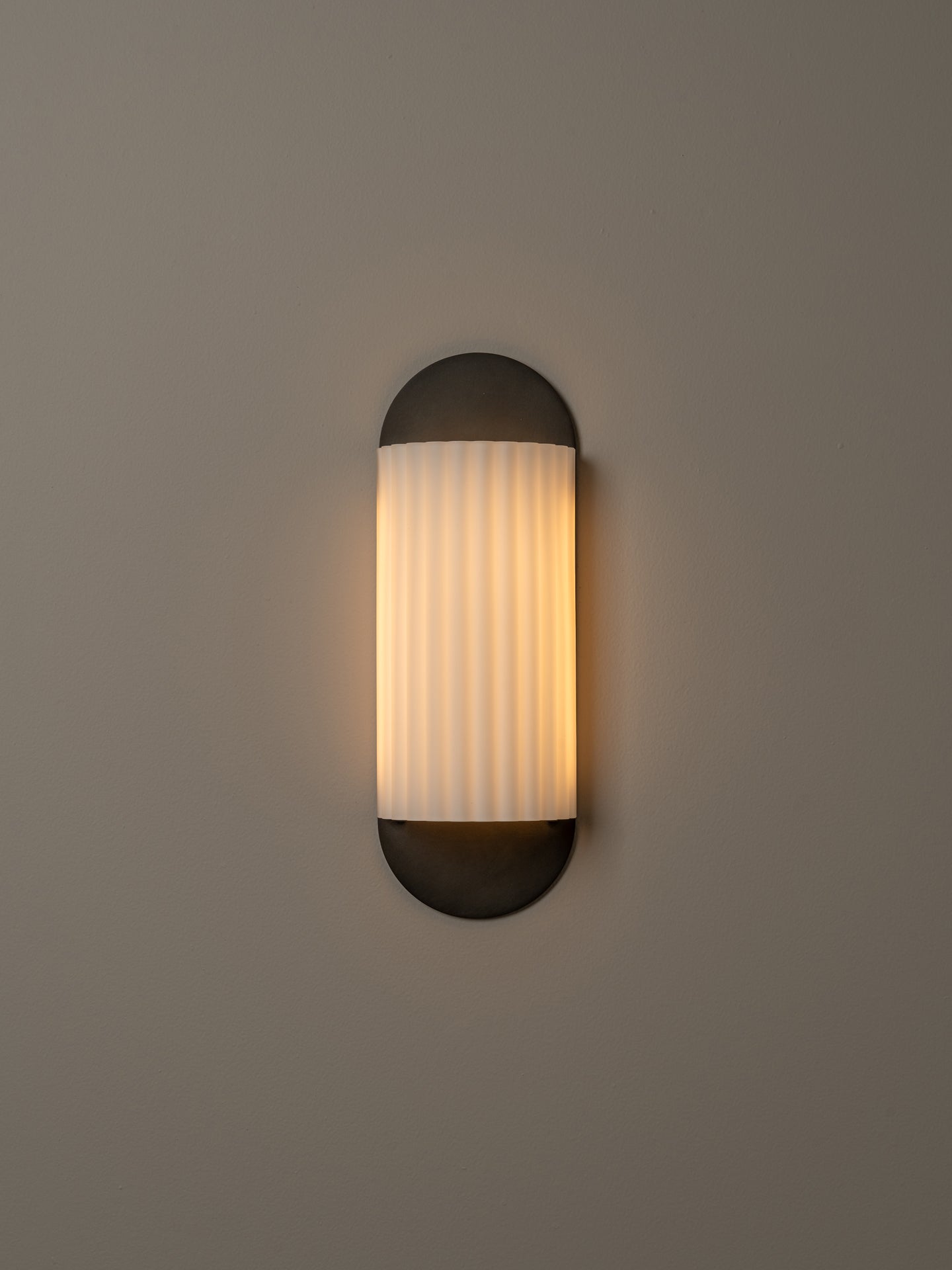 A modern wall sconce light fixture with a vertical ribbed frosted glass shade and dark metal end caps, mounted on a plain warm gray wall.