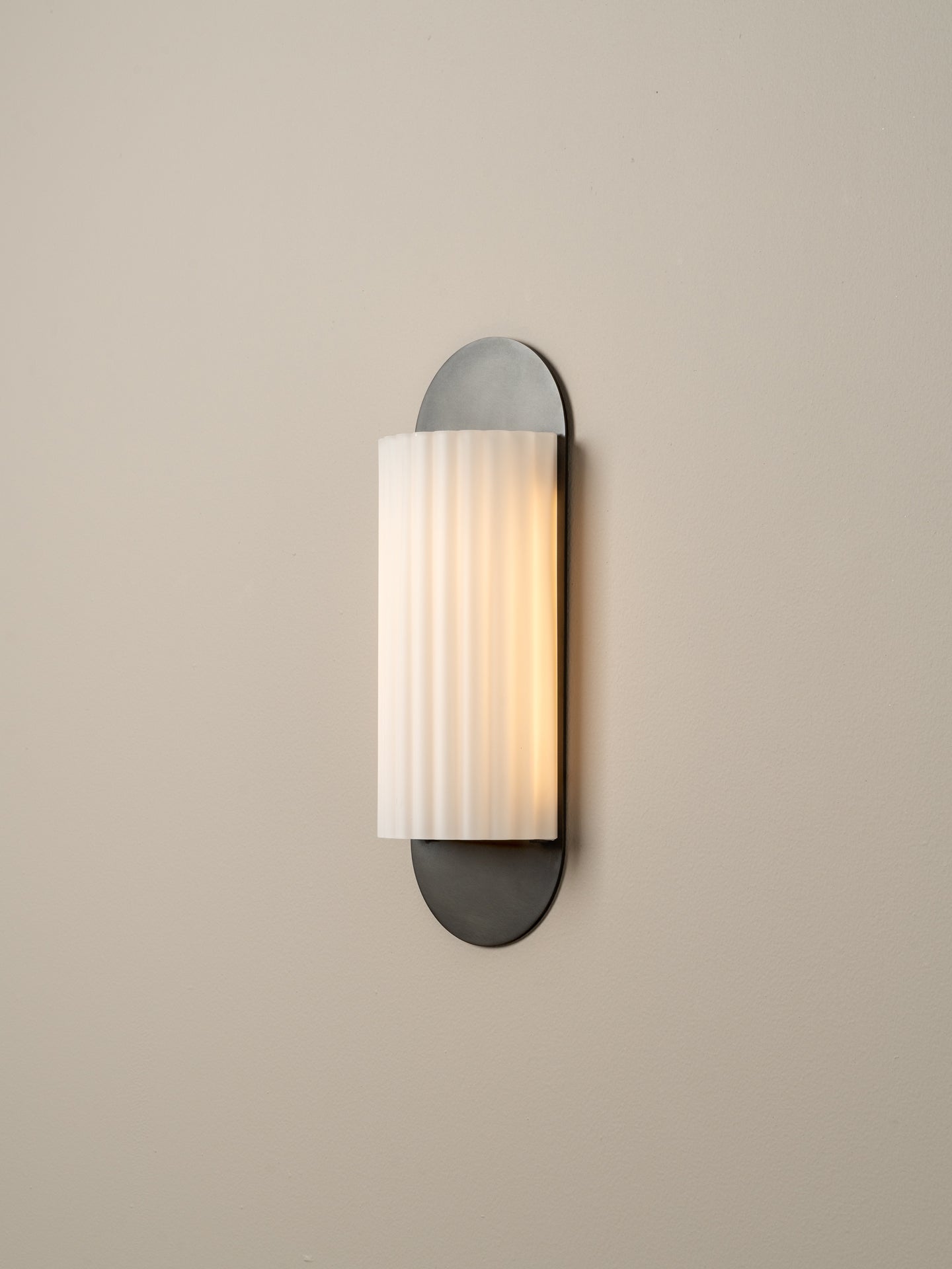 A modern wall sconce light fixture with a vertical ribbed white glass shade and dark gray metal mounting, shown illuminated against a plain beige wall.