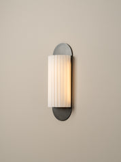 A modern wall sconce light fixture with a vertical ribbed white glass shade and dark gray metal mounting, shown illuminated against a plain beige wall.
