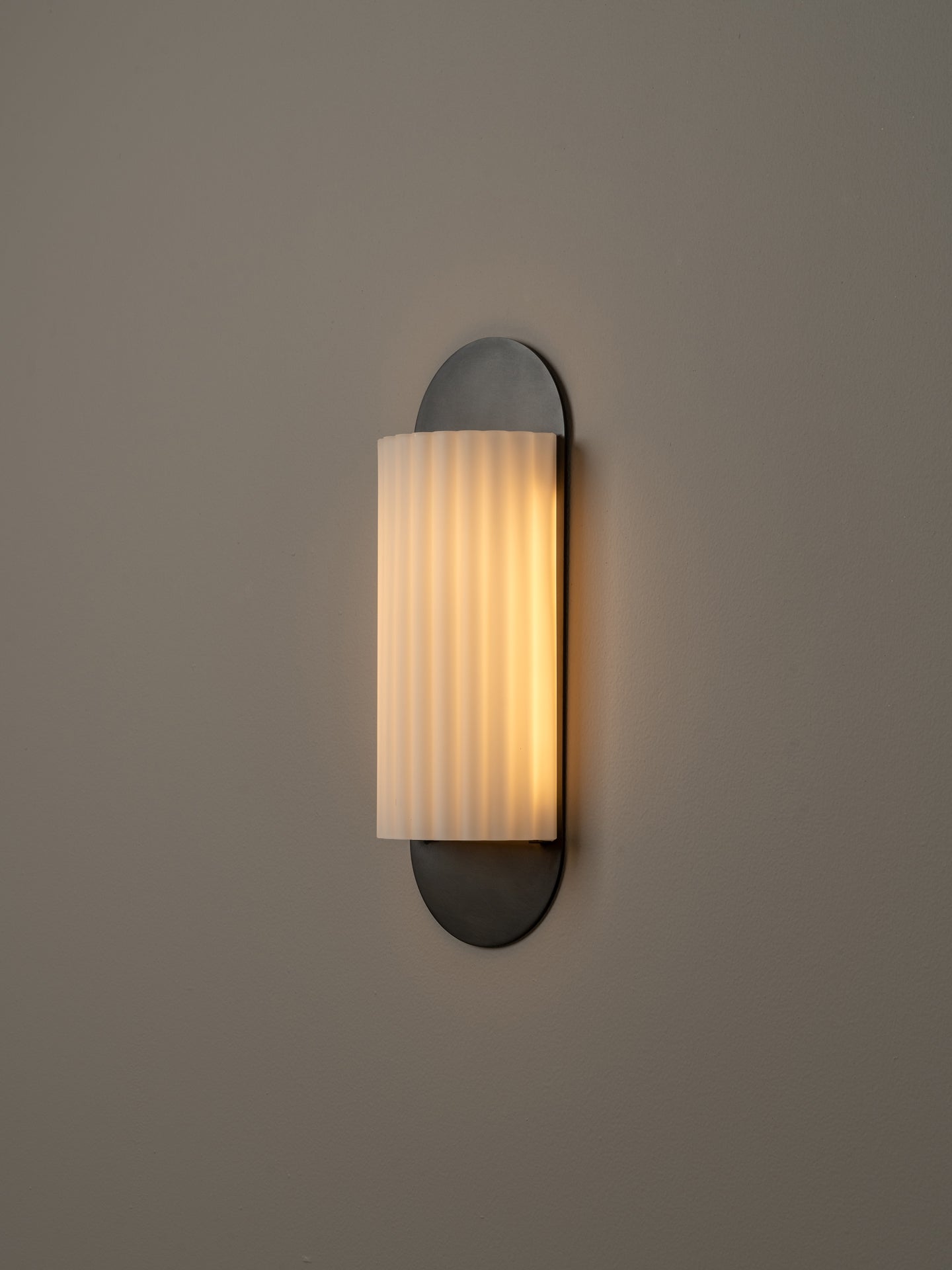 A modern wall-mounted sconce with a vertical pleated frosted glass shade and a dark metal base, shown lit against a plain warm gray wall.