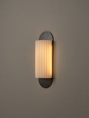 A modern wall-mounted sconce with a vertical pleated frosted glass shade and a dark metal base, shown lit against a plain warm gray wall.