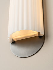 A close-up of a wall-mounted light fixture with a vertical ribbed frosted glass shade, a warm glowing rectangular diffuser, and a round brushed metal base against a plain wall.