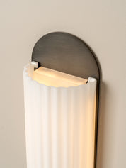 A modern wall sconce light fixture with a curved brushed metal backplate and a ribbed white glass shade, emitting a warm yellow light against a neutral beige wall.