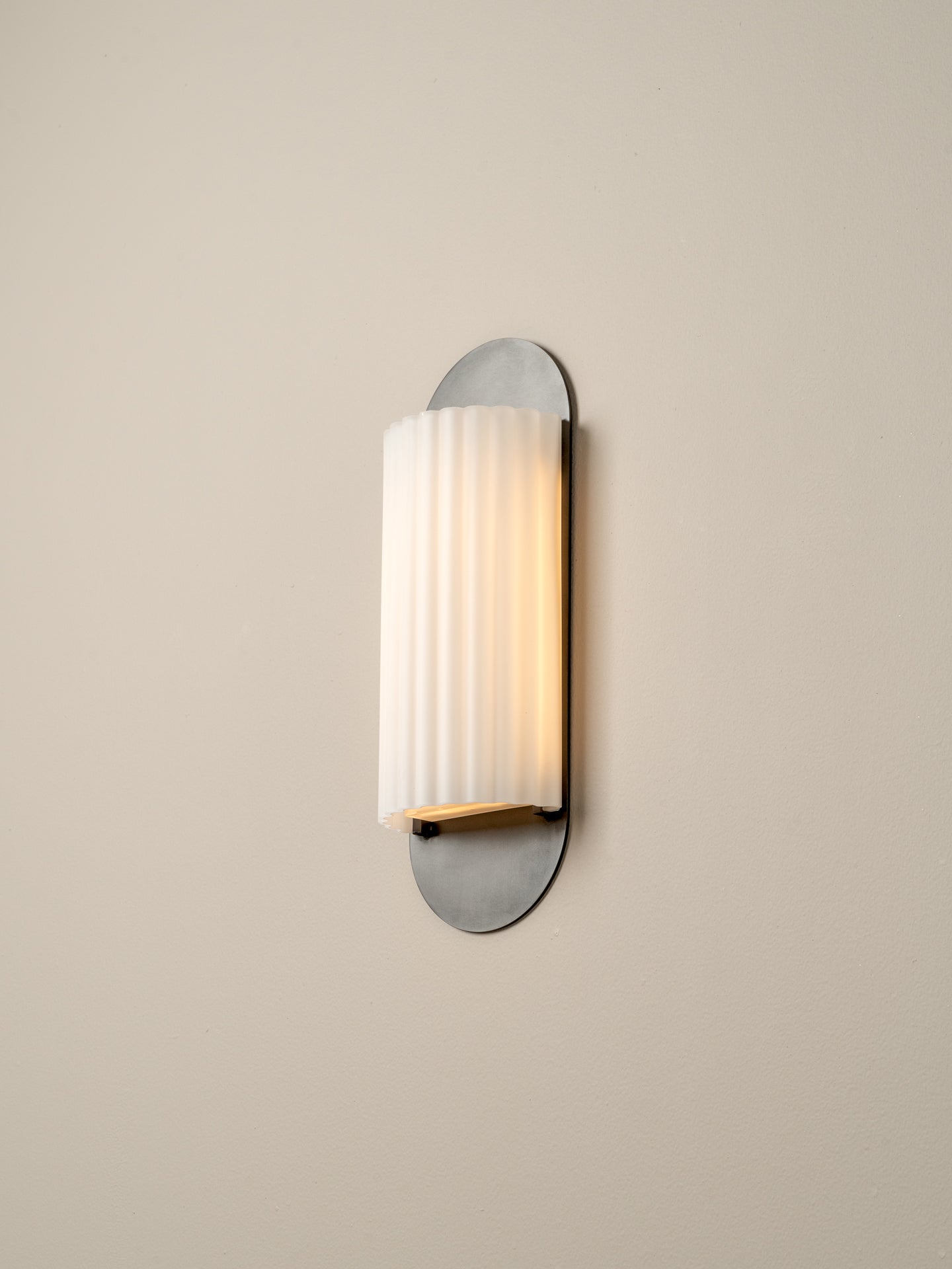 A modern wall-mounted sconce with a vertically fluted white glass shade and a brushed silver metal base, shown lit against a plain beige wall.