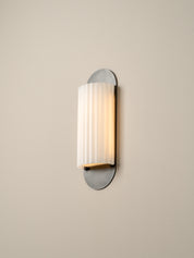 A modern wall-mounted sconce with a vertically fluted white glass shade and a brushed silver metal base, shown lit against a plain beige wall.
