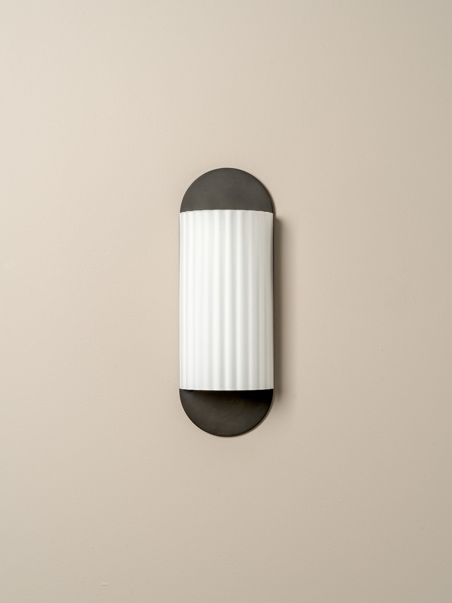 A modern wall-mounted light fixture with a vertically oriented cylindrical white ribbed shade and dark charcoal metal caps at the top and bottom, shown against a plain beige wall.