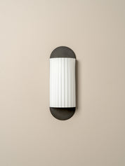 A modern wall-mounted light fixture with a vertically oriented cylindrical white ribbed shade and dark charcoal metal caps at the top and bottom, shown against a plain beige wall.