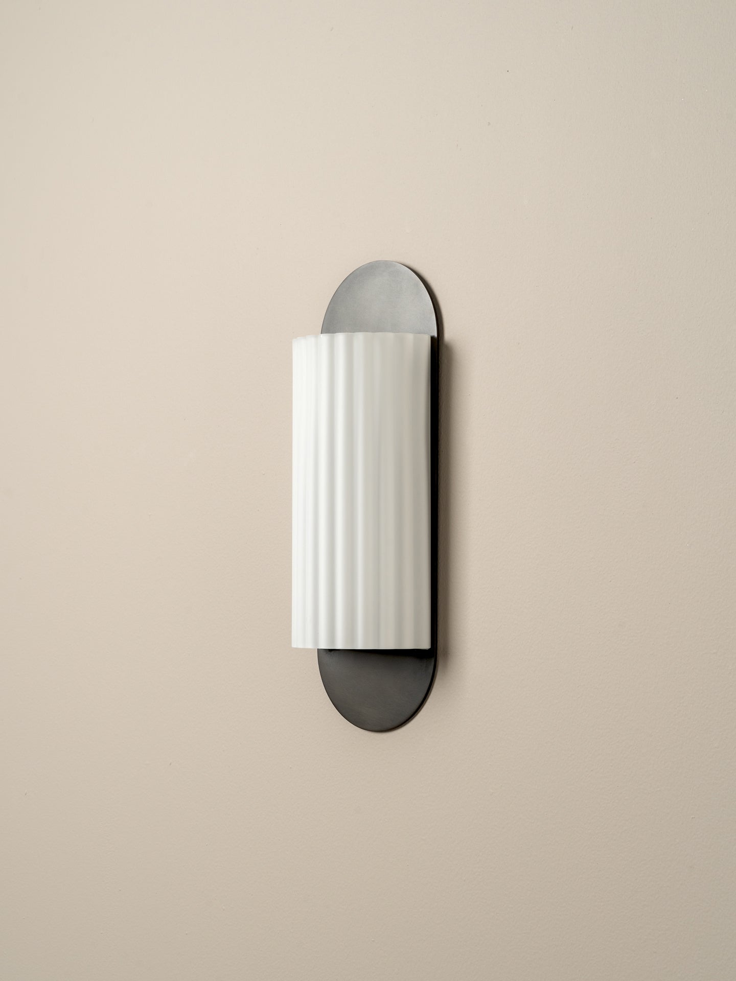 A modern wall-mounted sconce with a vertically ribbed white cylindrical shade and dark gray metal base and backplate, shown against a plain beige wall.