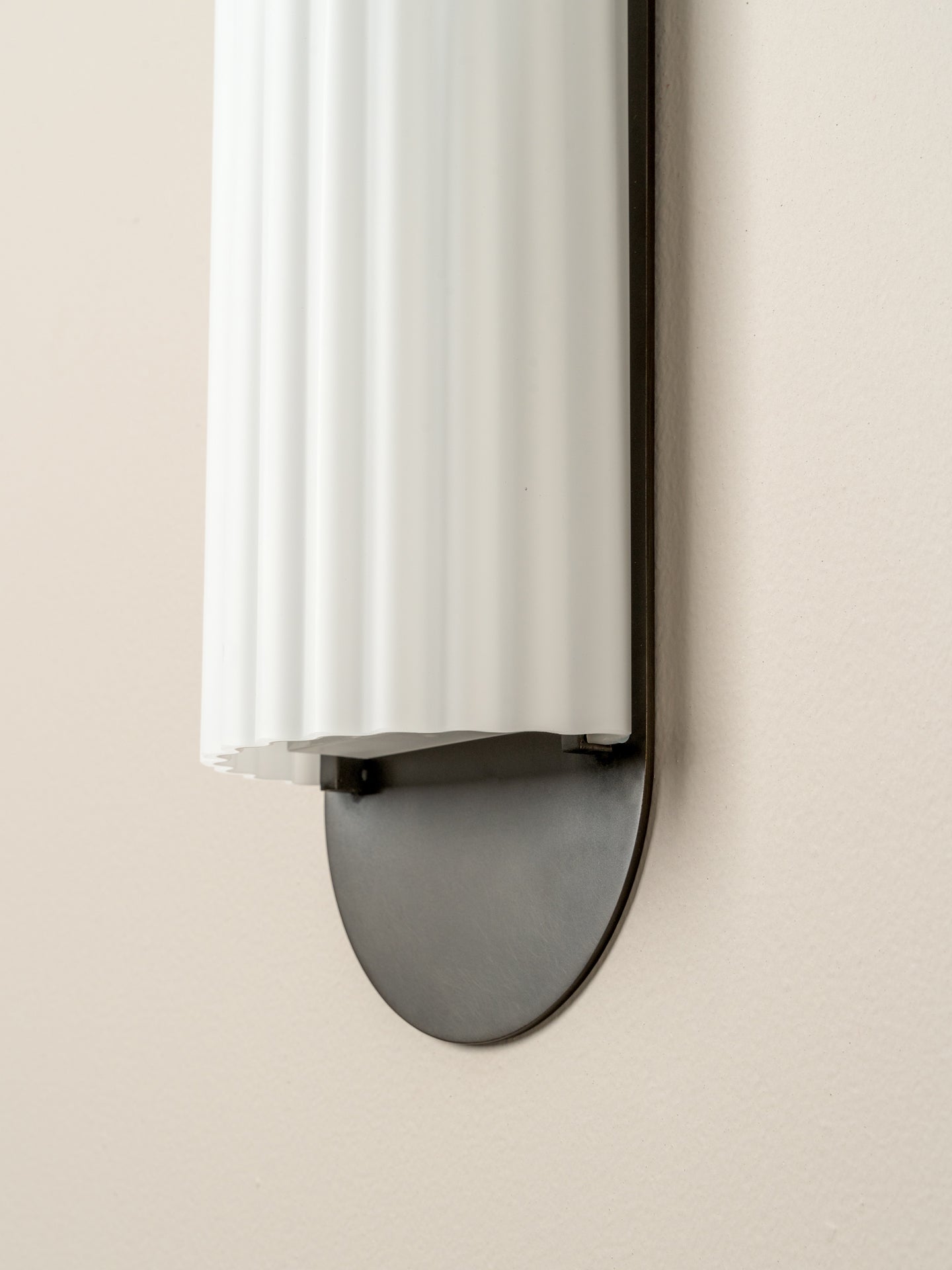 A modern wall-mounted light fixture with a white ribbed cylindrical shade and a dark charcoal metal base, shown mounted on a warm beige wall.