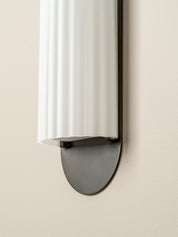 A modern wall-mounted light fixture with a white ribbed cylindrical shade and a dark charcoal metal base, shown mounted on a warm beige wall.