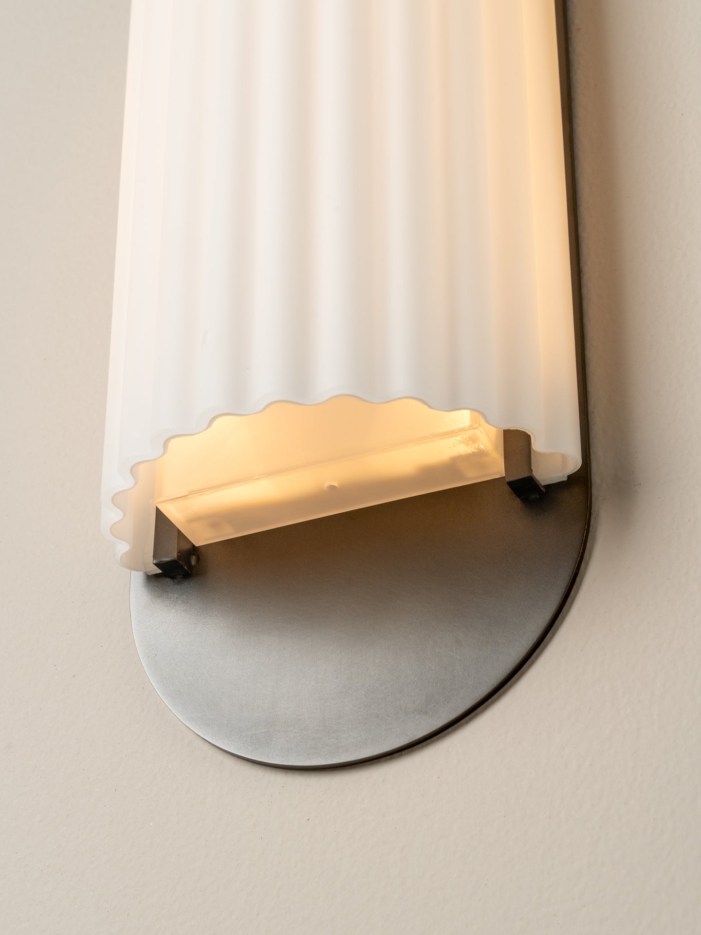 A close-up of a wall-mounted light fixture with a vertical ribbed frosted glass shade, a warm glowing rectangular diffuser, and a round brushed metal base against a plain wall.