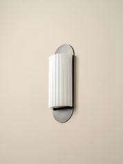 A modern wall-mounted sconce with a white ribbed cylindrical shade and brushed silver metal mounting plates, shown against a plain beige wall.